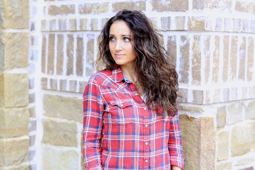 Woman with curly hair wearing a red plaid shirt standing outside in front of a brick background