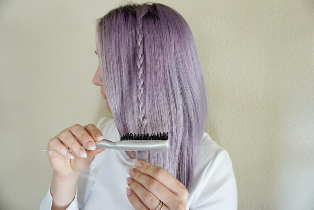 Woman with lavender dyed hair wearing a white shirt uses a brush to comb her hair