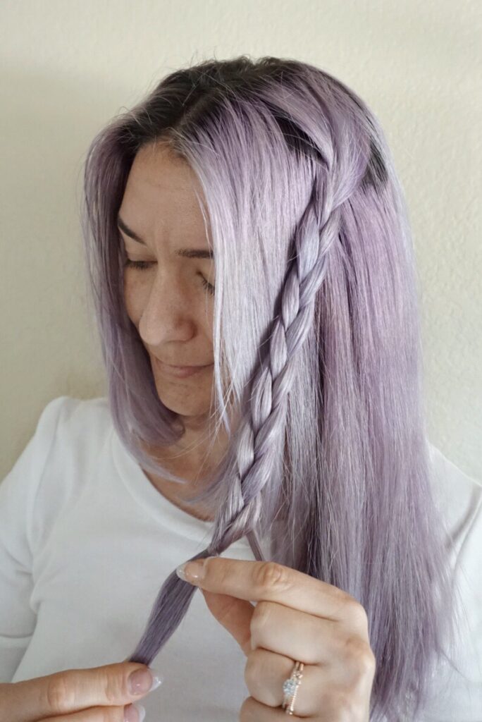 Woman with lavender dyed hair wearing a white shirt braids her hair