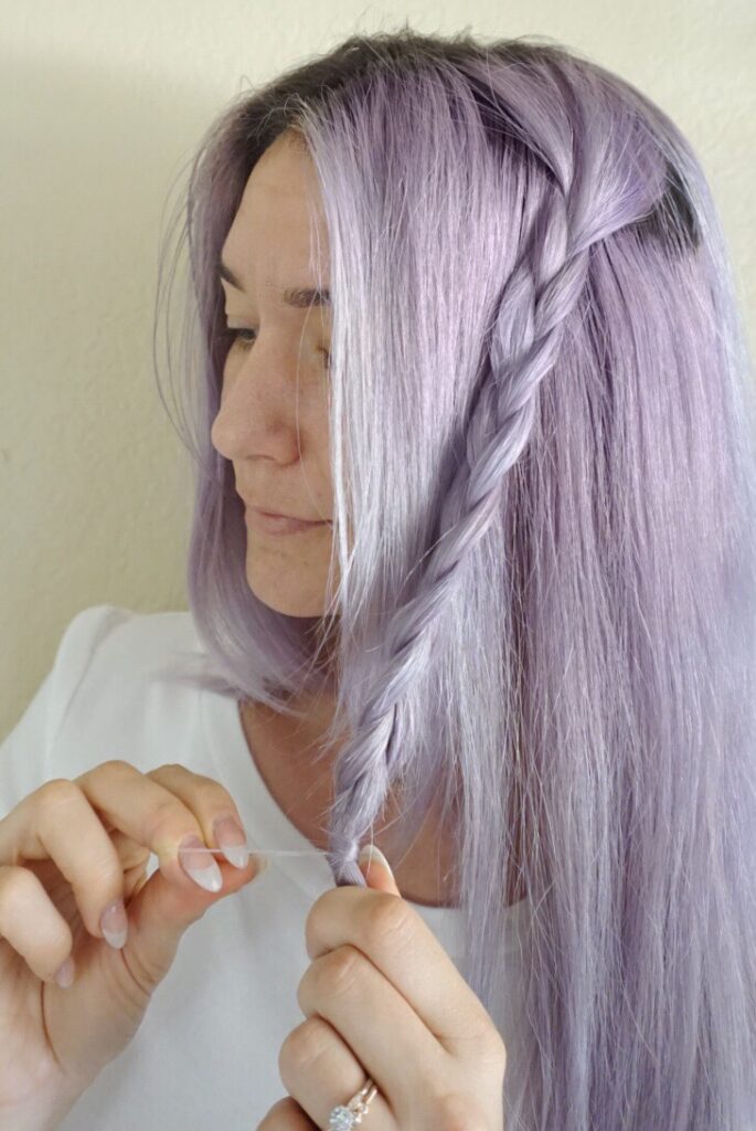 Woman with lavender dyed hair wearing a white shirt uses a hair tie to secure her braid