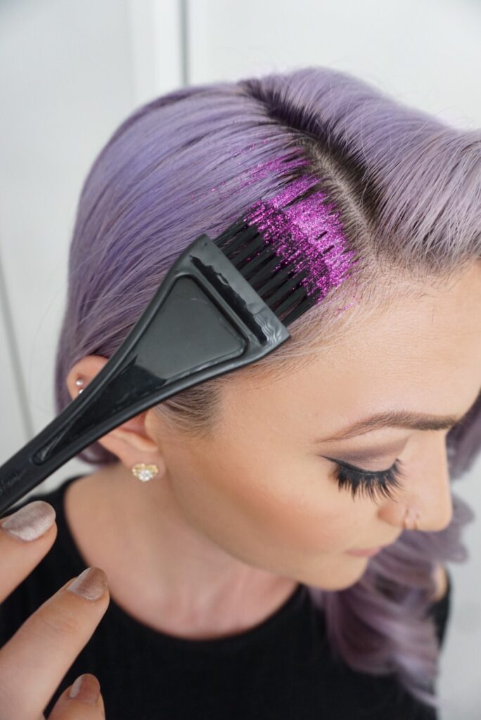Woman uses styling brush to apply purple glitter gel to her lavender colored hair