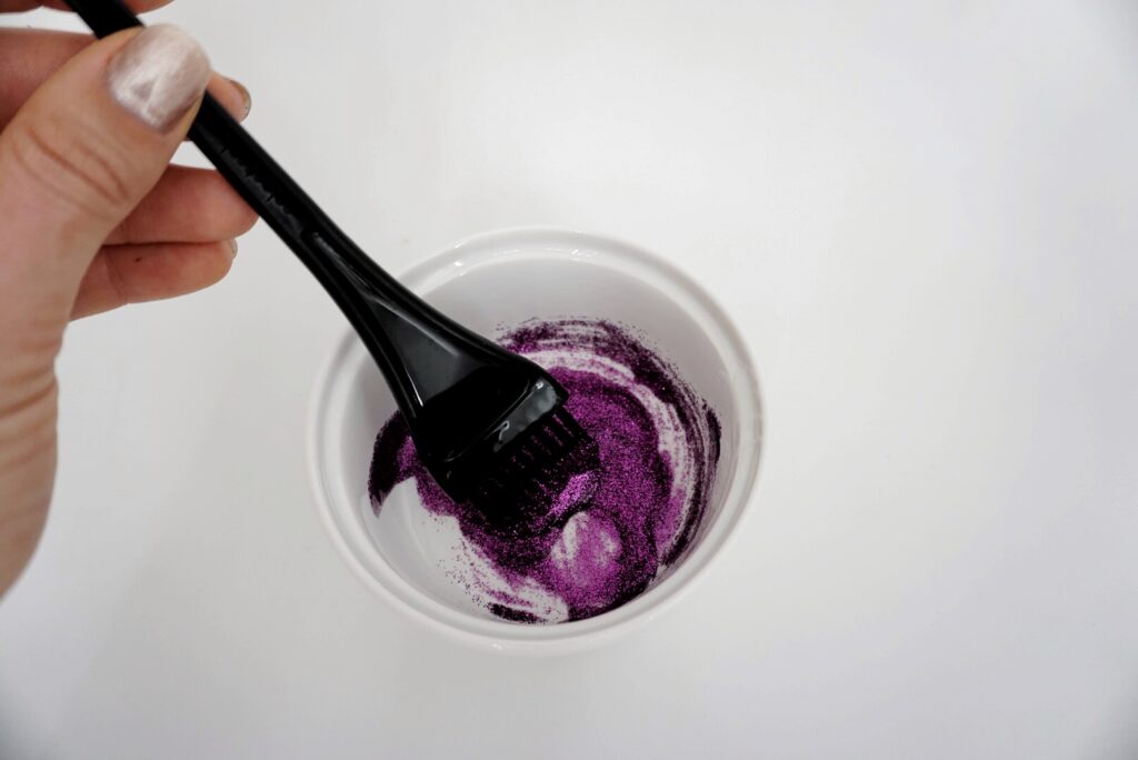 Woman mixing hair gel and purple glitter in a white bowl with a styling brush 