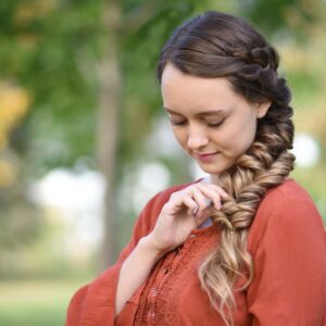 Girl outside wearing an orange shirt modeling "Side Elastic Braid" hairstyle and touching her hair