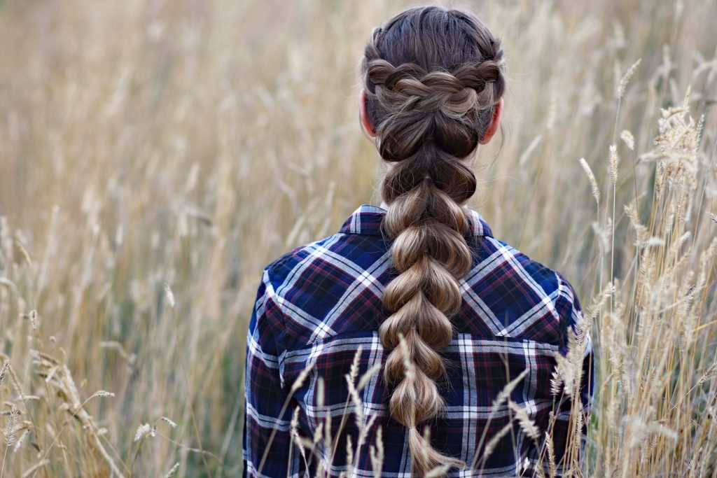 Back view of young girl in the meadow wearing blue plaid shirt modeling "Wrapped Pull-Thru Braid" hairstyle