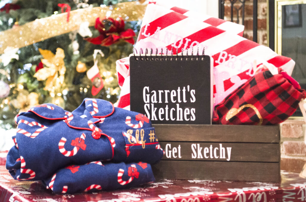 Black notebook, a plaid bag, and red and white blanket placed inside of a brown box, sitting next to blue Christmas pajamas close to a Christmas tree