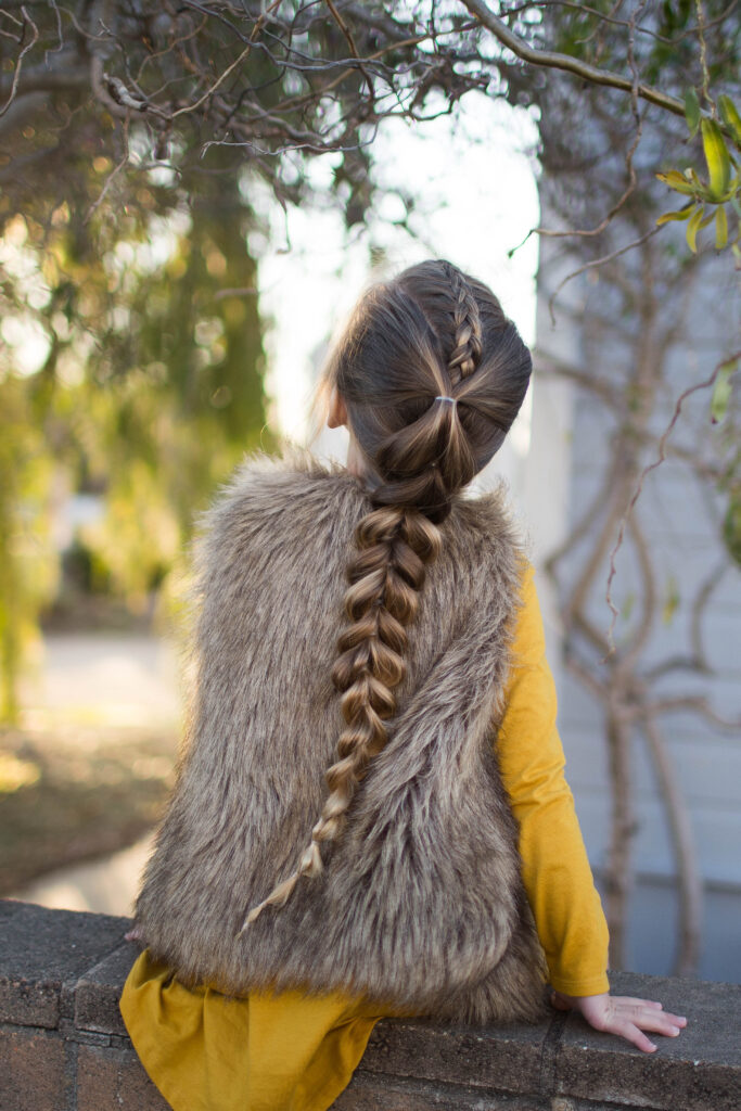 Back view of girl wearing faux fur vest over yellow dress outside modeling the "Dutch Pull-Thru Combo Braid" hairstyle