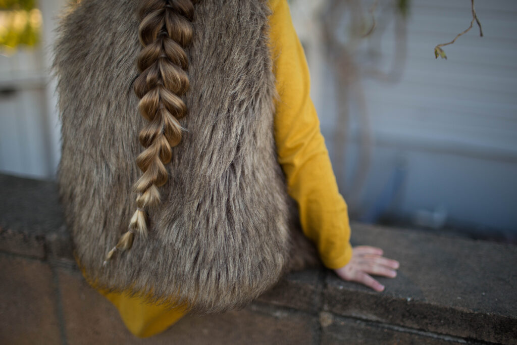 back view a little girl with long braided hair wearing a faux fur vest over a yellow dress