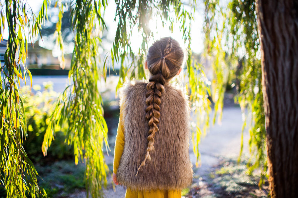 Back view of girl wearing faux fur vest over yellow dress outside modeling the "Dutch Pull-Thru Combo Braid" hairstyle