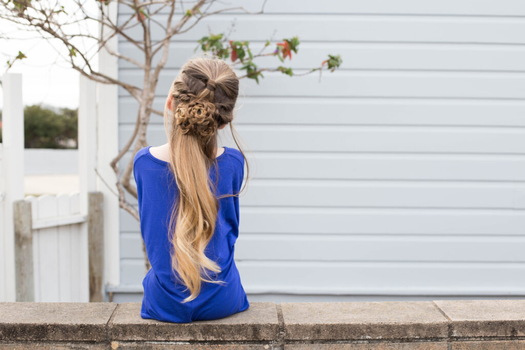 Back view of a little girl wearing a blue dress sitting outside modeling "Flower Half-Up" hairstyle