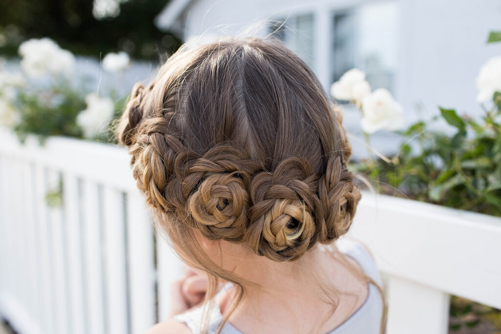 Back view of young girl standing outside in front of white flowers modeling "Flower Crown Braid" hairstyle