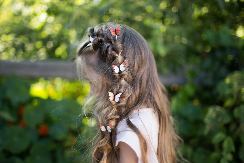 Side view of a young girl with hair standing outside modeling "Flip Faux Fishtail" hairstyle with butterfly accessories