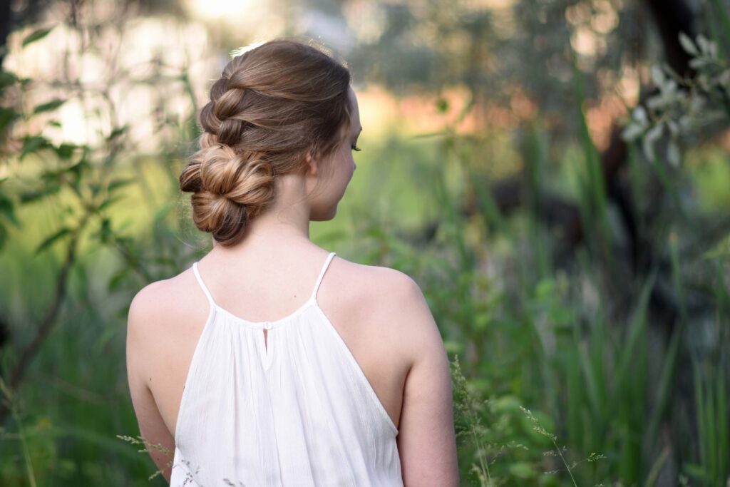 Side view of girl with a white dress standing outside in greenery and modeling "Knotted Braid Updo" hairstyle.