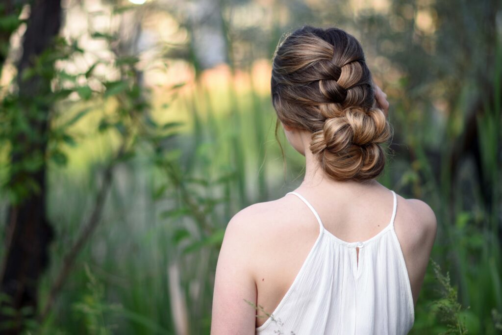 Backside view of girl with a white dress standing outside in greenery and modeling "Knotted Braid Updo" hairstyle.