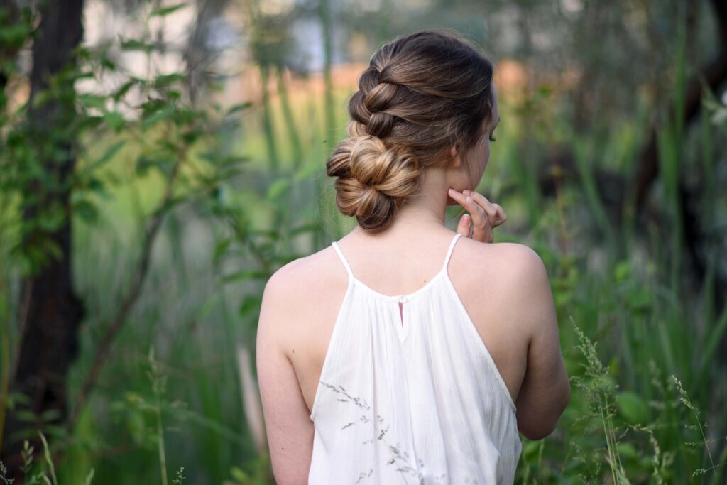 Side view of girl with a white dress standing outside in greenery and modeling "Knotted Braid Updo" hairstyle.