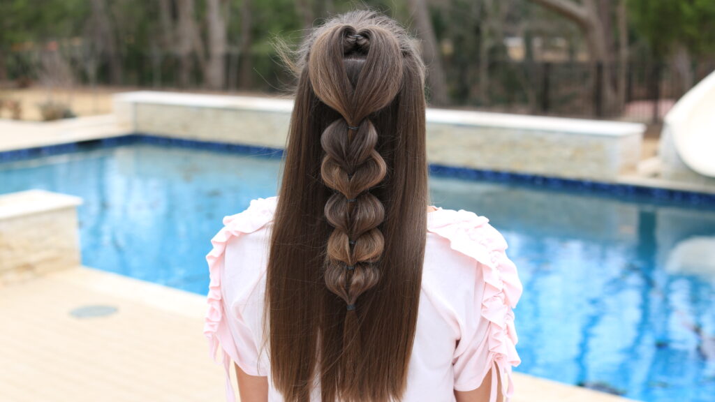 Back view of girl standing outside in front of the pool modeling the "Heart Pull-Thru Braid" hairstyle