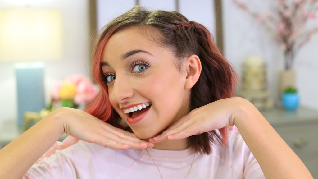 Girl with pink dyed hair indoors holding hands to her chin modeling "Side Pull Back" hairstyle.