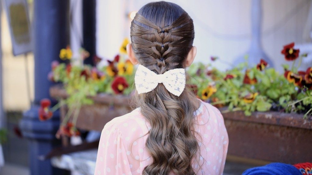 Back view of girl with pink shirt standing outside in front of flower bed modeling "Mermaid Heart Braid" hairstyle with a bow in her hair.