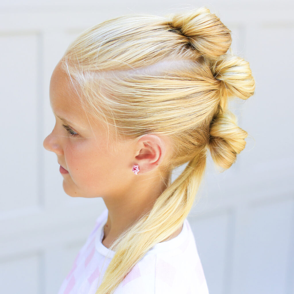 Side view of blonde girl standing outside in front of a white background modeling "Triple Bow Hawk" hairstyle