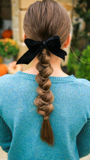 Girl standing outside with a blue shirt modeling "Wrapped Braid" hairstyle with a black bow