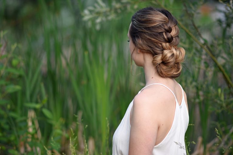 Backside view of girl with a white dress standing outside in greenery and modeling "Knotted Braid Updo" hairstyle.