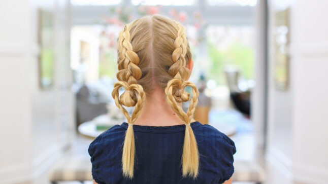 Back view of blonde girl standing indoors modeling the "Dutch Heart 2 in 1" hairstyle with double braids.