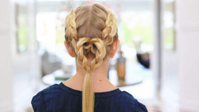 Back view of blonde girl standing indoors modeling the "Dutch Heart 2 in 1" hairstyle with single braid