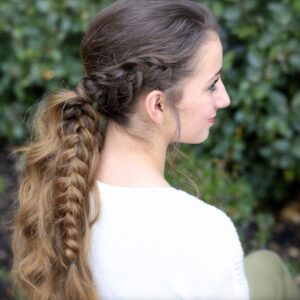 Side profile image of smiling girl standing in front of outside bushes modeling a "Viking Braid"